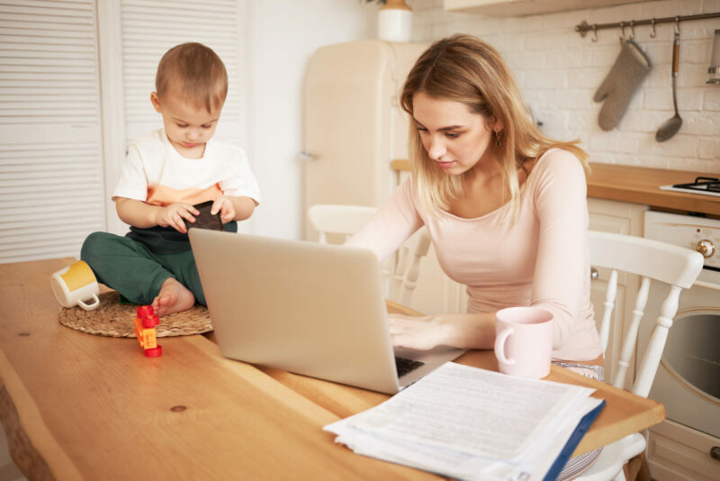 Worried upset young blonde female sitting at kitchen table with papers and portable computer feeling stressed because she has to make report and take care of her baby son while he is staying at home